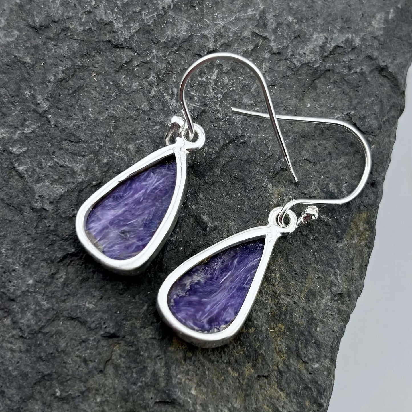 Purple Charoite teardrop earrings with silver hooks on a dark stone surface