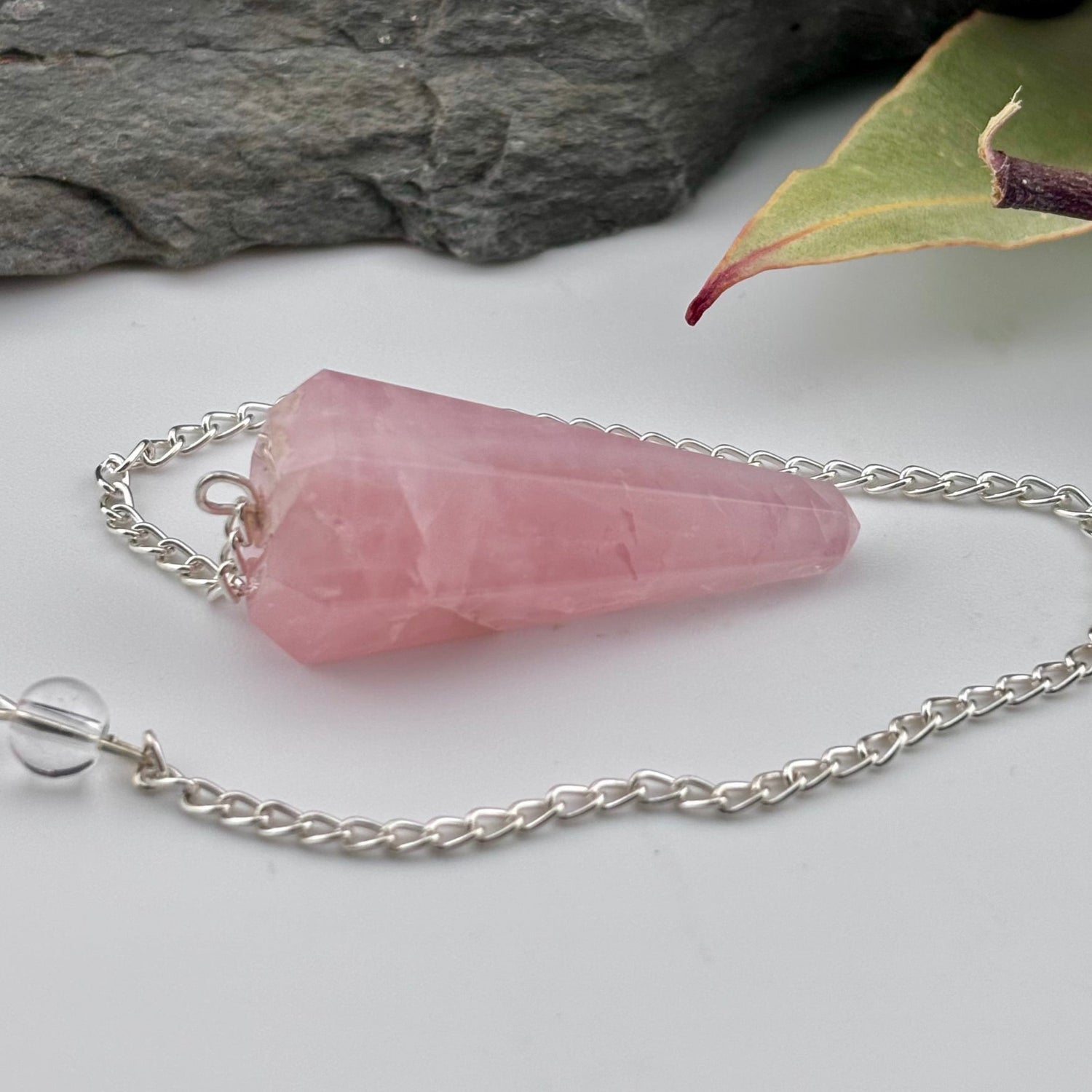 Pink crystal pendant on a silver chain with a stone and leaf in the background