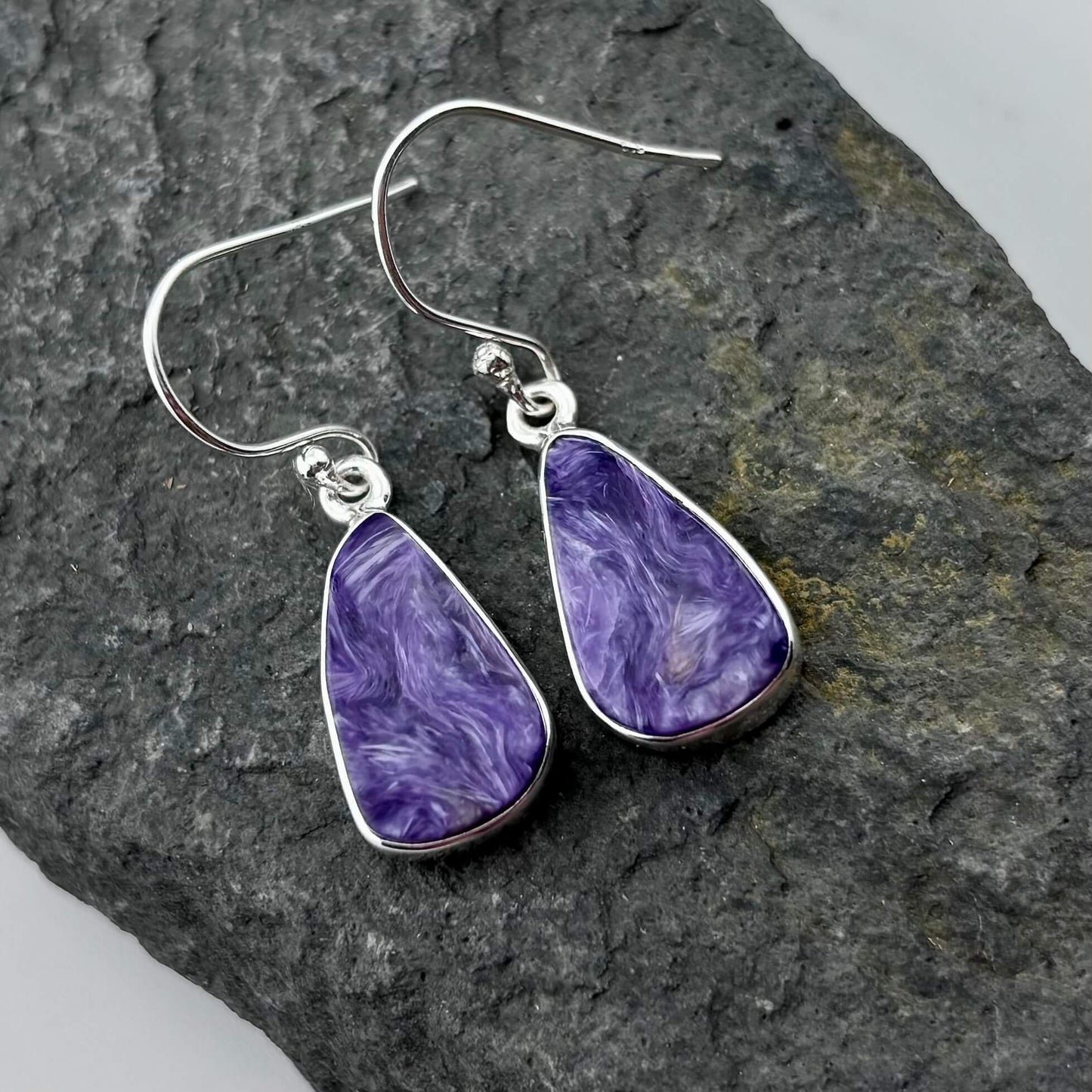 Purple Charoite teardrop earrings with silver hooks on a dark stone surface