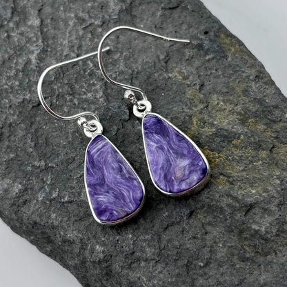 Purple Charoite teardrop earrings with silver hooks on a dark stone surface