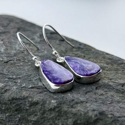 Purple Charoite teardrop earrings with silver hooks on a dark stone surface