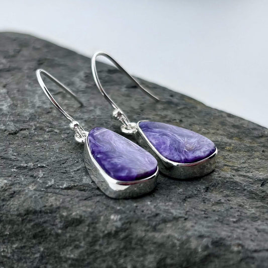 Purple Charoite teardrop earrings with silver hooks on a dark stone surface