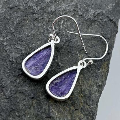 Purple Charoite teardrop earrings with silver hooks on a dark stone surface